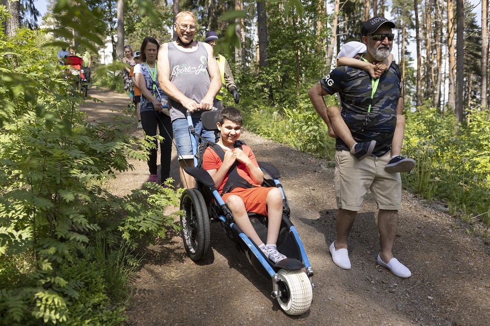 perheet metsäretkellä. Isoisä työntää poikaa maastopyörätuolissa ja vieressä kävelee isä jolla reppuselässä pikkuveli