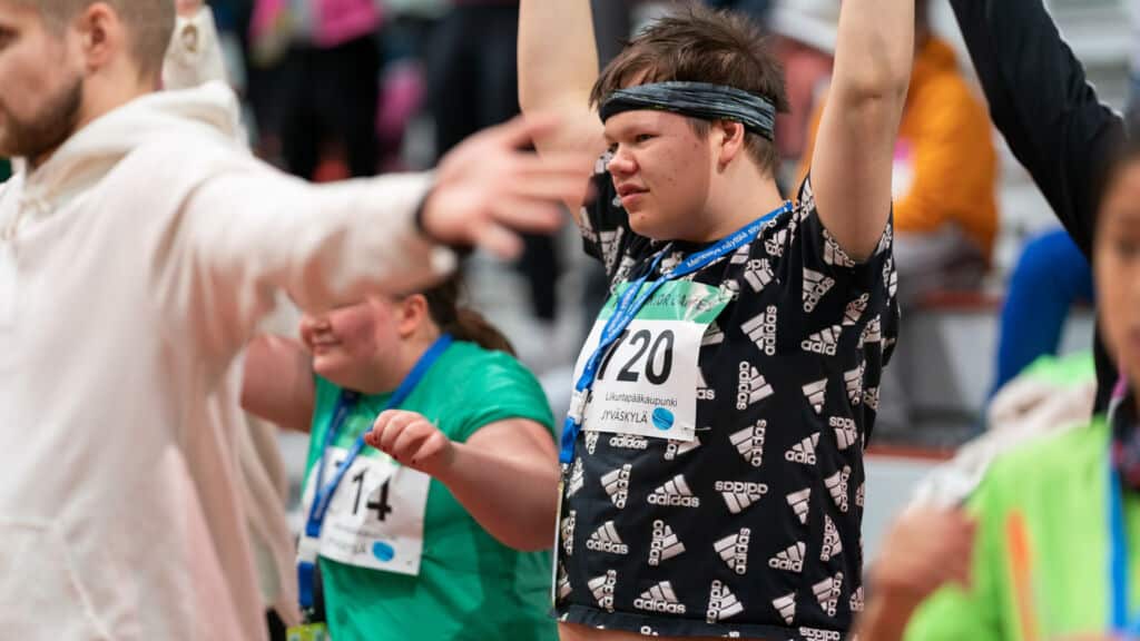 Kuva urheilukilpailuista. Kuvassa nuori mies tuulettaa kädet ilmassa ja hänellä on kaulassaan voitettu mitali.