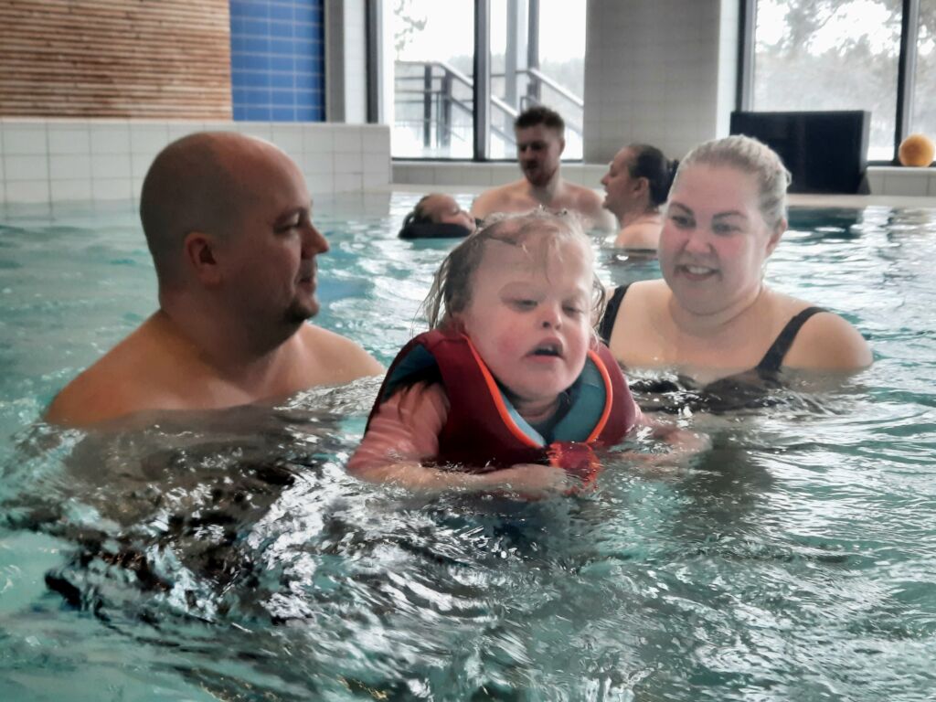 Kuvassa perhe on uimassa uimahallissa. Perheeseen kuuluu isä, äiti ja lapsi. He ovat iloisia.