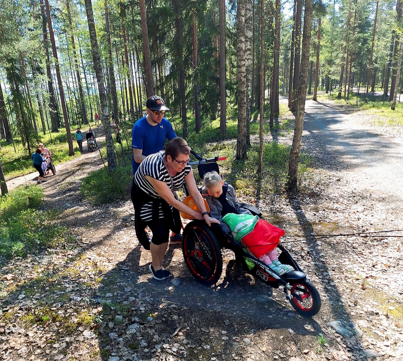 Kaksi aikuista työntää maastopyörätuolissa istuvaa lasta ylämäkeen metsäpolkua pitkin. Pyörätuolin vetonarut ovat kireällä, vaikkei vetäjä näy kuvassa. Taustalla näkyy ihmisiä ja maastopyörätuoleja.