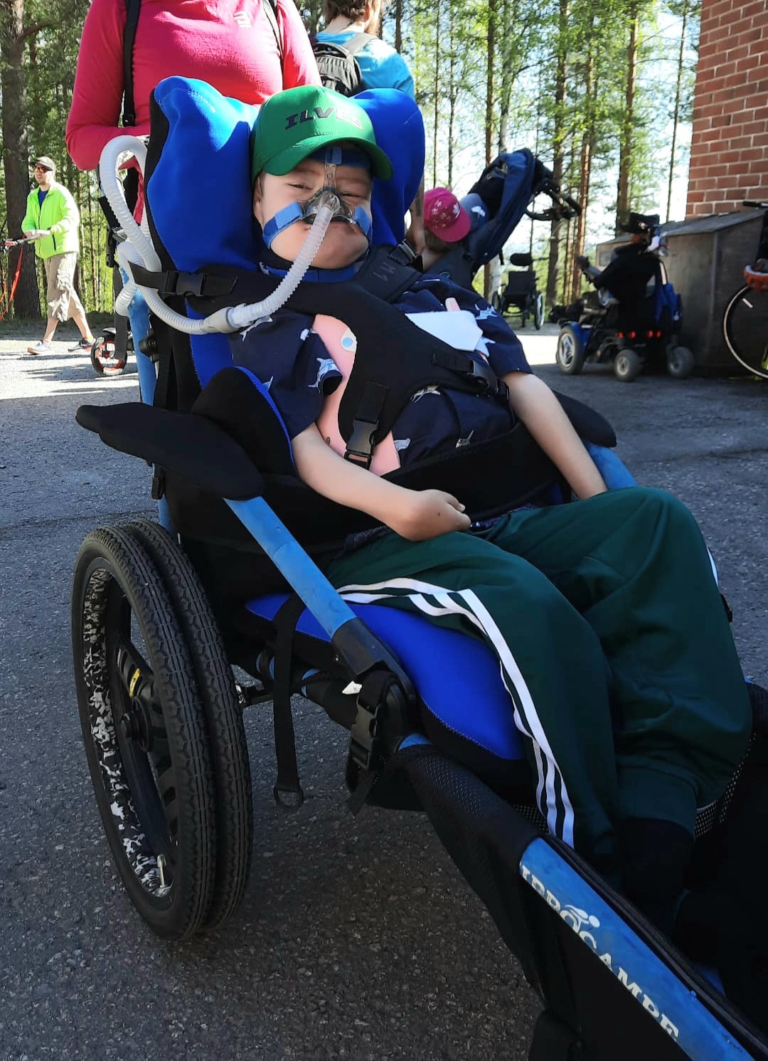 Poika istuu sinisessä maastopyörätuolissa. Pyörätuoliin on aseteltu sininen asentotyyny. Pojalla on hengitystuki.