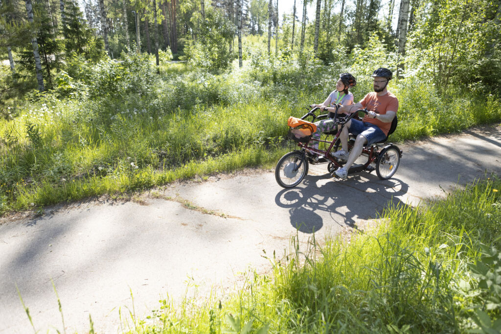 Mies ja tyttö polkevat rinnakkain poljettavalla pyörällä vehreän kesäisessä ympäristössä.