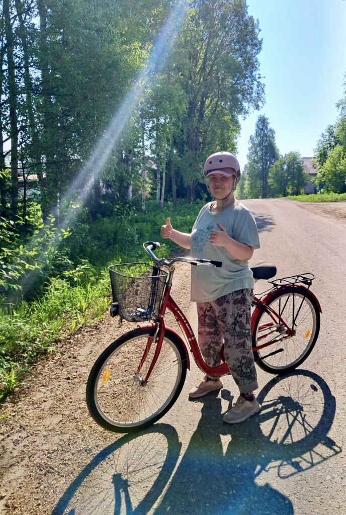 Nainen on polkupyörän selässä. Taustalla on metsää ja aurinko paistaa.
