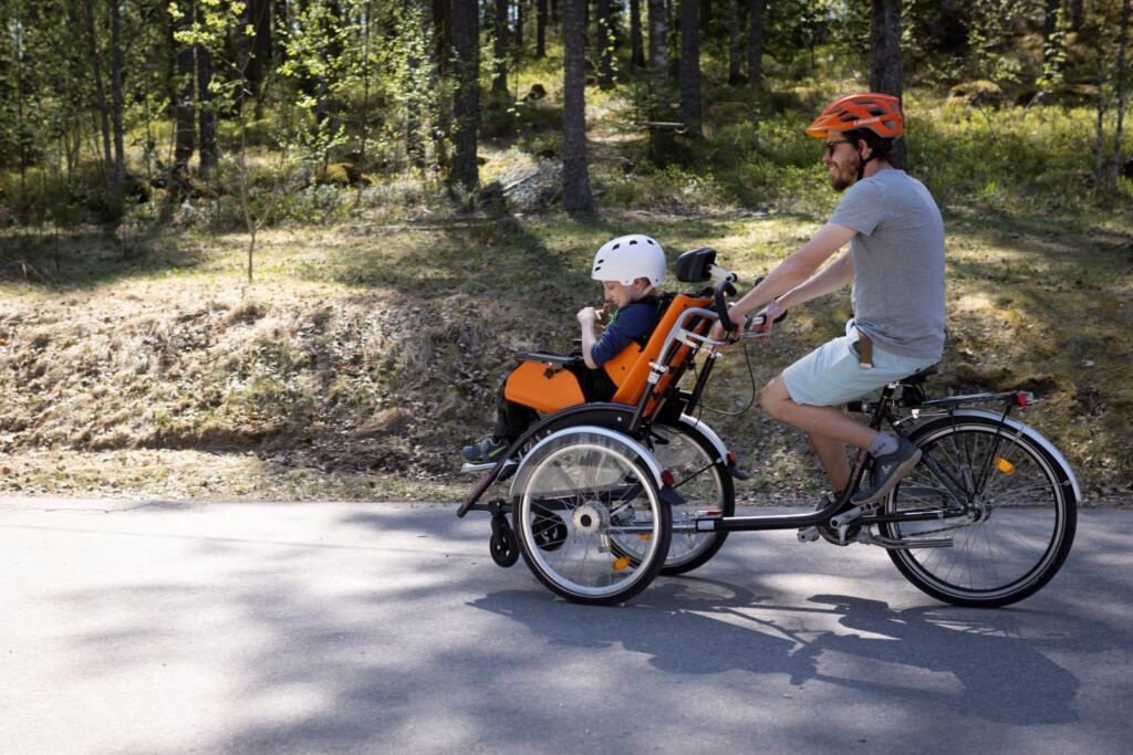 Mies ja poika pyöräilevät pyörätuolinkuljetuspyörällä aurinkoisessa säässä.