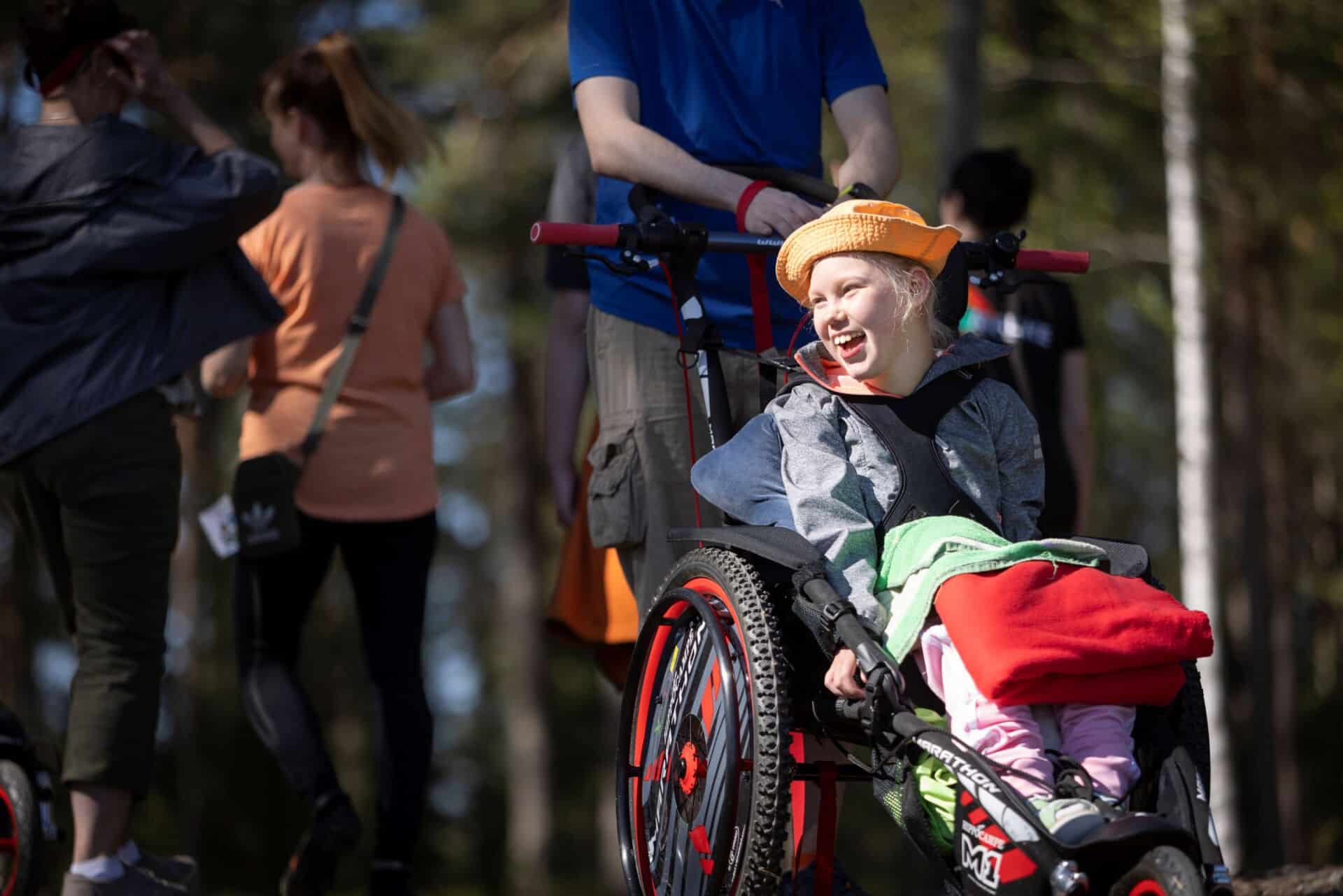 Aurinko paistaa. Tyttö istuu maastopyörätuolissa, jota t-paitaan pukeutunut aikuinen työntää.