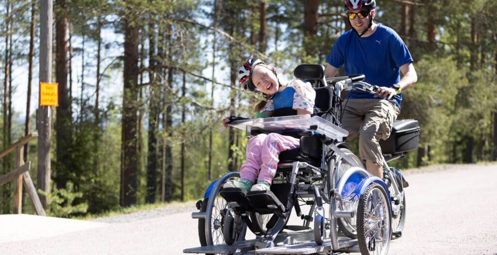 Lapsi ja aikuinen pyöräilevät yhdistelmäpyörällä, jonka etuosassa on pyörätuolimainen istuin. Lapsi istuu siinä. Aikuinen polkee ja ajaa pyörän takaosassa.