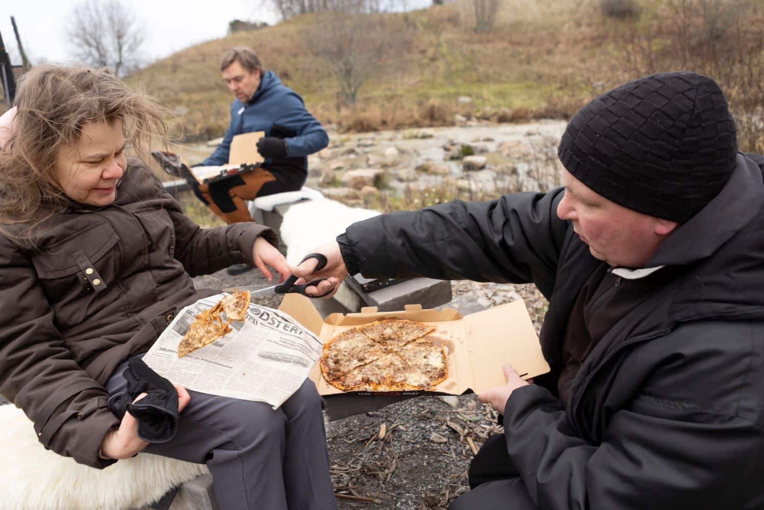 Kolme henkilöä istuu ulkona retkipaikalla. Etualalla mies antaa naiselle pizzaa ja leikkaa sitä saksilla. Taka-alalla istuu mies pizzalaatikko sylissään.