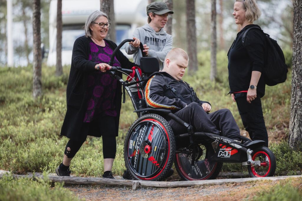 Mies maastopyörätuolissa metsäpolulla. Kaksi ihmsitä työntää pyörätuolia takaa ja yksi seisoo vieressä kasvot työntäjiin päin.