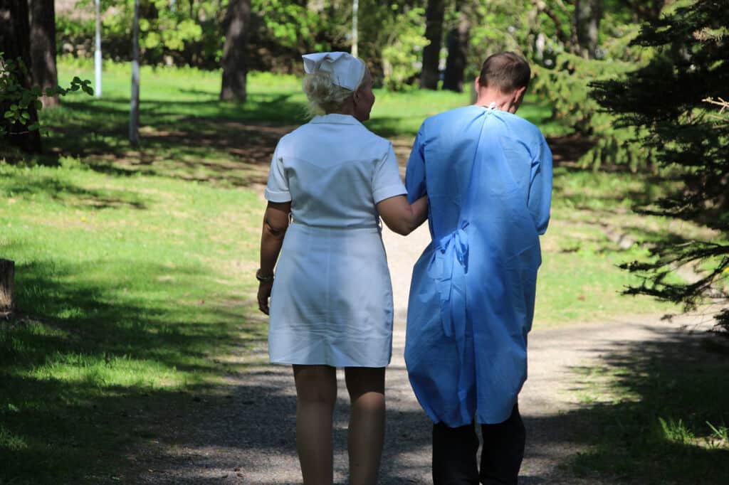 Kuvassa on kaksi ihmistä selkä kameraan päin. He kävelevät käsikynkässä ulkona. Toisella on sairaalavatteet päällä ja toisella sairaanhoitajan vaatteet.