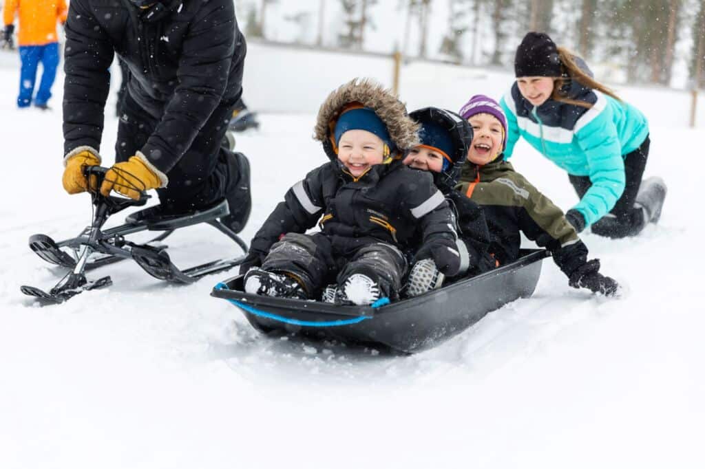 Kolme pientä lasta laskee mäkeä pulkalla. Takana aikuinen työntää pulkalle vauhtia. Vieressä toinen aikuinen laskee rattikelkalla.