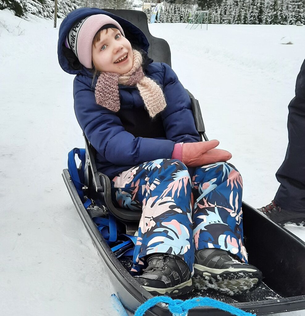 On talvi. Lapsi istuu mustassa ahkiossa, johon on kiinnitetty korkeaselkänojainen penkki.