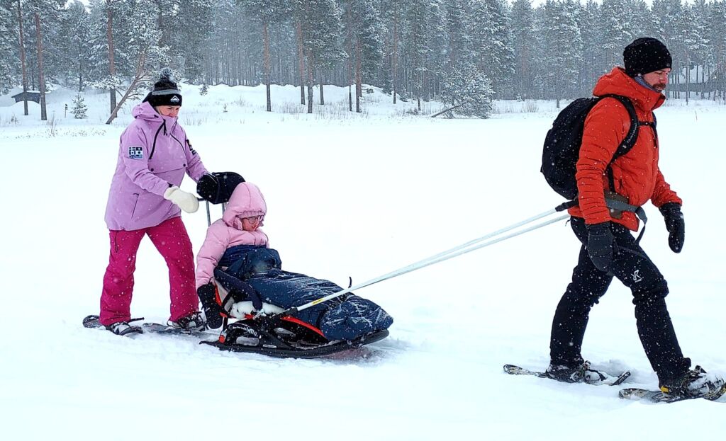 Lapsi istuu Snow Comfort -kelkassa järven jäällä. Taustalla näkyy mäntymetsää. Nainen työntää kelkkaa työntökahvoista ja mies vetää vetoaisoista. Heillä on lumikengät jalassaan. Kelkassa on istuinosa, jossa lapsi mahtuu istumaan jalat suorana ja korkea selkä-/niskatuki.