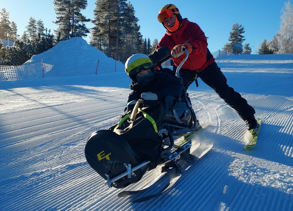 Kelkkalaskettelija ja avustaja laskevat alas laskettelurinnettä. Kelkassa on kaksi suksea.