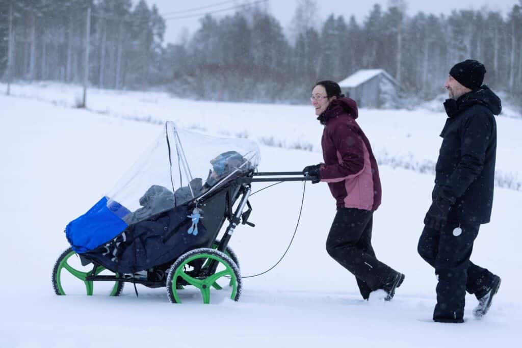 Maassa on paljon lunta. Lapsi istuu monitoimirattaissa, joita nainen työntää lumihangessa.