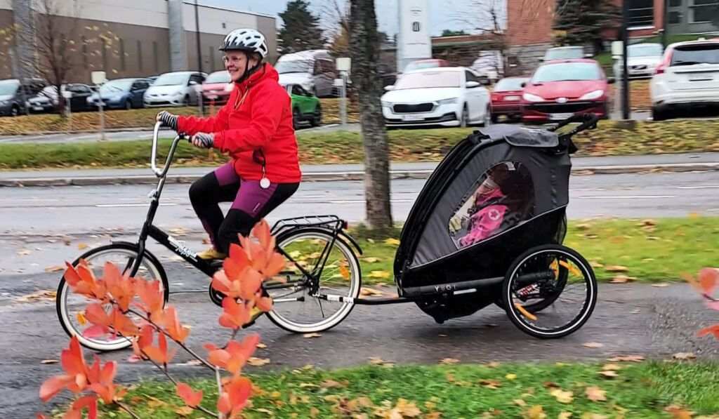 Nainen pyöräilee pyörällä, jonka perässä on musta kuomullinen peräkärry. Kärryssä istuu lapsi.