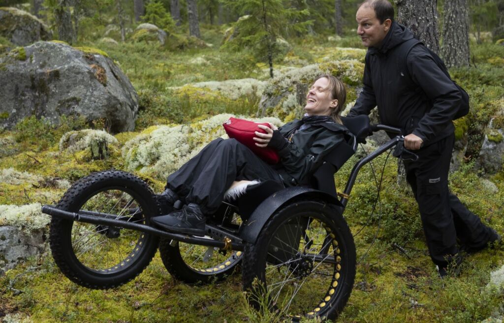 Nainen istuu maastopyörätuolissa keskellä metsää. Maasto on sammeleista ja kivikkoista. Maastopyörätuolia työntää mies. Mies painaa pyörätuolin työntökahvaa alas, niin että tuolin eturengas on ilmassa.